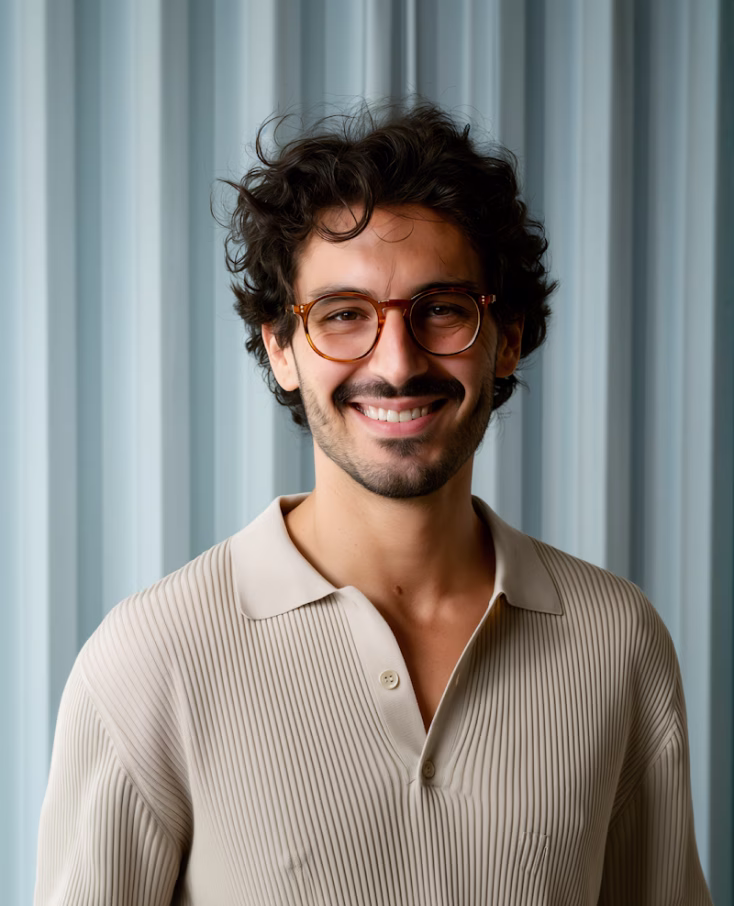 Smiling man with curly dark hair, beard, and glasses wearing a beige ribbed polo shirt against a light blue curtain background.