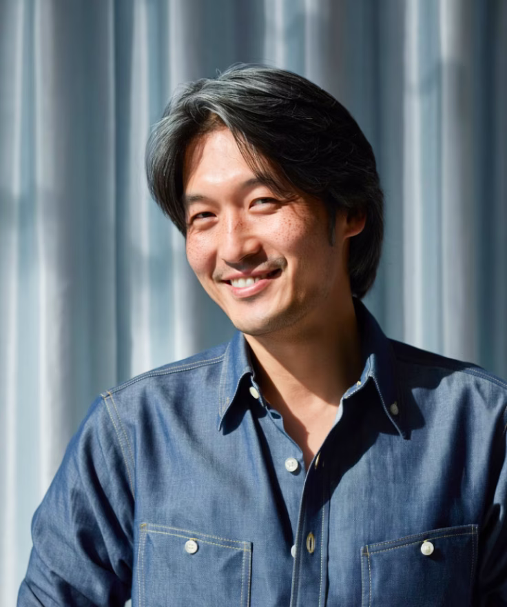 Smiling man with medium-length black hair wearing a blue denim shirt standing in front of a curtain.