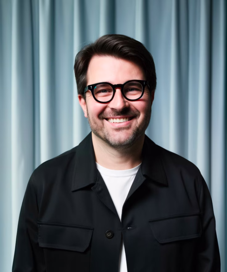 Smiling man with dark hair, beard, and round black glasses wearing a black jacket and white shirt against a blue curtain background.