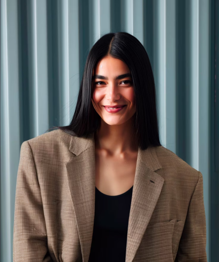 Young woman with long black hair wearing a brown checkered blazer and black top, smiling in front of a corrugated metal wall.