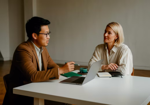 Two colleagues having a discussion at a white table with a laptop, notebook, and coffee cup.